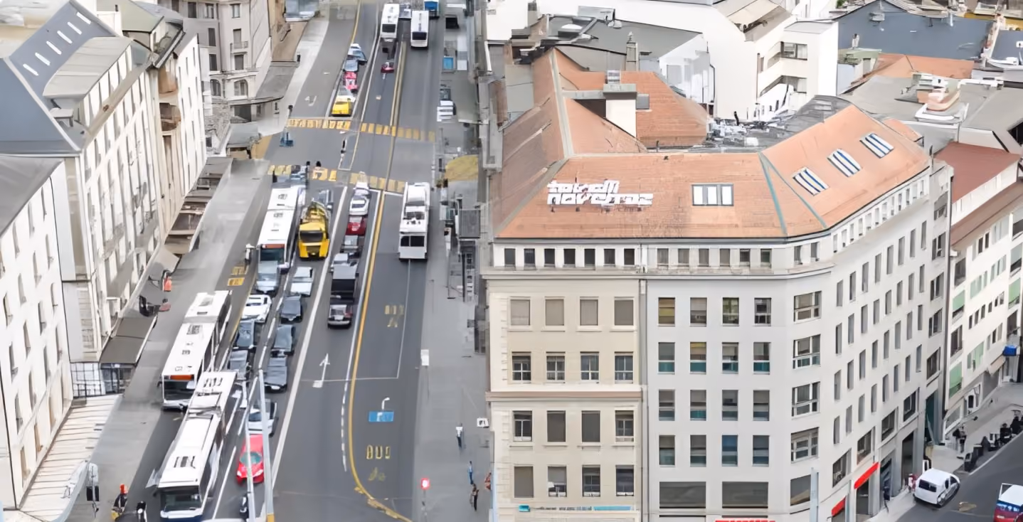 Une vue en hauteur d'une rue animée en ville montrant une circulation dense, avec plusieurs bus et voitures alignés à un croisement à côté d'un grand bâtiment à plusieurs étages avec un toit en pente marron et une enseigne blanche sur le toit.