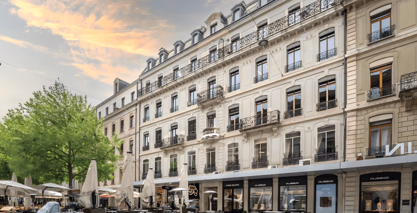 Un élégant bâtiment de plusieurs étages sur la Place du Molard à Genève sous un coucher de soleil, avec des boutiques de luxe au rez-de-chaussée et la terrasse d'un café avec des parasols fermés au premier plan.