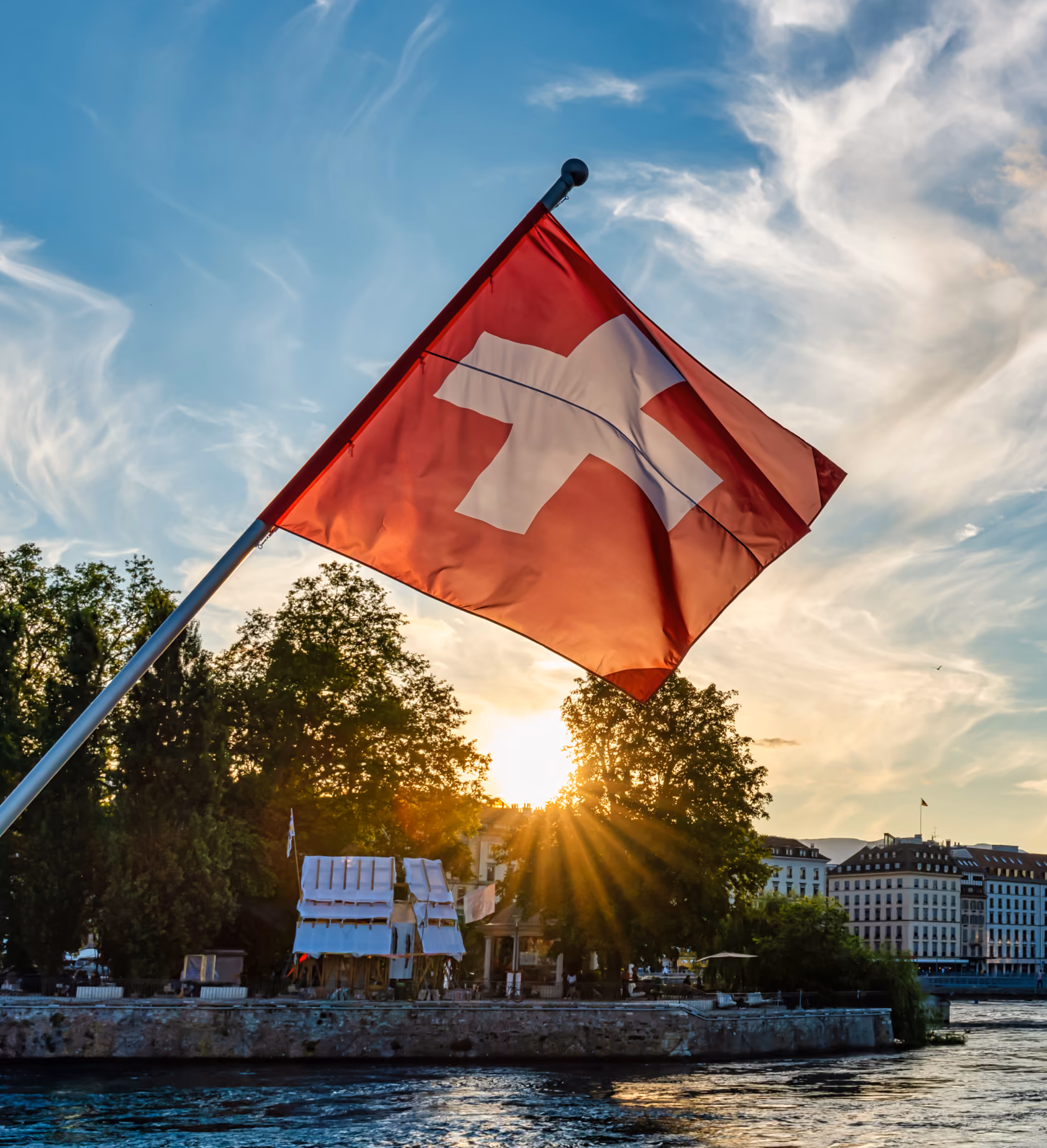 Un drapeau suisse flottant sur un bateau au coucher du soleil, avec les rayons du soleil perçant à travers les arbres le long de la rive genevoise en arrière-plan.