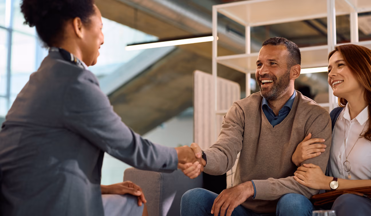 Un homme et une femme souriants sont assis ensemble, l'homme serrant la main d'une femme professionnelle assise en face d'eux dans un bureau moderne et lumineux.