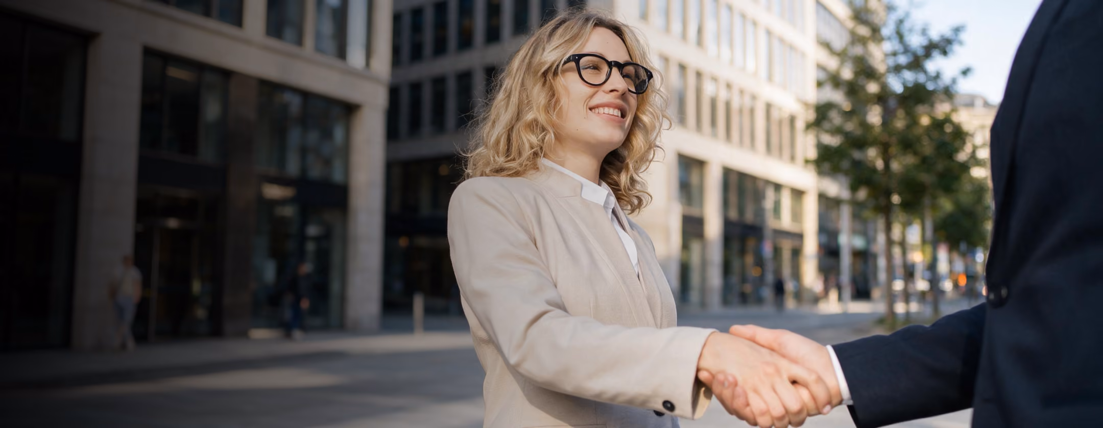 Une femme d'affaires blonde souriante en blazer beige et lunettes serre la main de quelqu'un dans une rue ensoleillée bordée de bâtiments modernes.