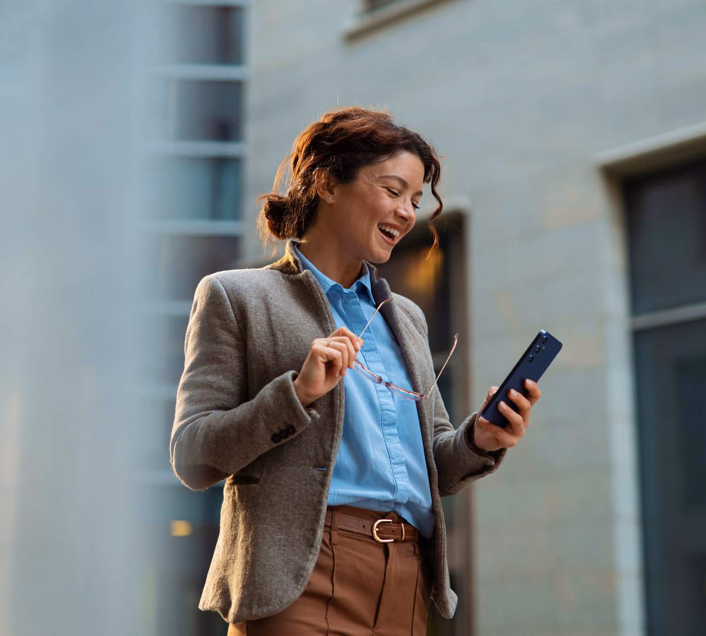 Une femme d'affaires souriante tenant ses lunettes tout en regardant son smartphone à l'extérieur devant un immeuble de bureaux moderne.