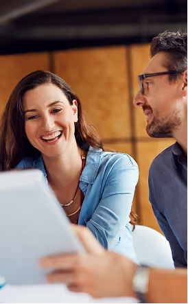 Une femme souriante en chemise en jean et un homme portant des lunettes regardant une tablette ensemble.