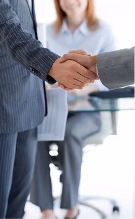 Deux professionnels des affaires se serrant la main au-dessus d'un bureau, avec une collègue souriante en arrière-plan.