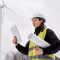 Une ingénieure portant un casque de sécurité blanc et un gilet haute visibilité tenant des plans et regardant une tablette près d'une éolienne.