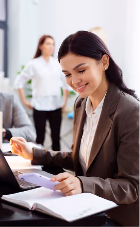 Une femme d'affaires souriante à son bureau tenant un smartphone au-dessus d'un carnet ouvert à côté d'un ordinateur portable.