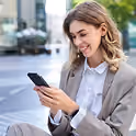 Une jeune femme d'affaires souriante en costume beige envoyant un message sur son smartphone à l'extérieur.