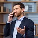 Un homme d'affaires barbu en veste de costume sombre faisant un geste de la main tout en parlant au téléphone portable dans un bureau avec des étagères en bois en arrière-plan.