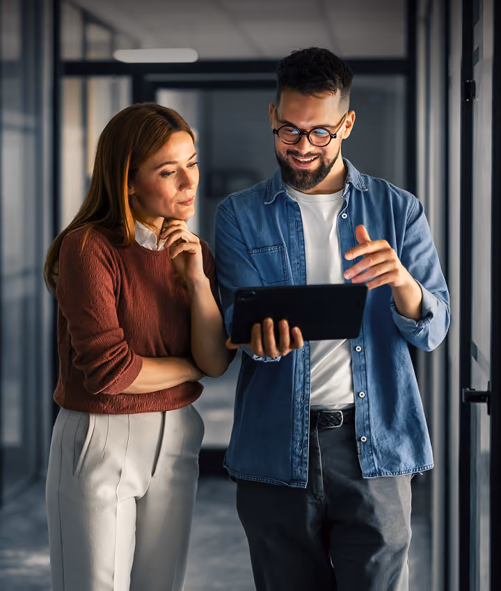 Un homme à lunettes en chemise en jean montre une tablette à une collègue attentive dans un couloir de bureau vitré.