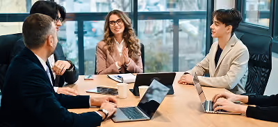 Un groupe de cinq professionnels est assis autour d'une table de conférence en bois dans un bureau moderne, souriants et participant à une réunion de travail avec des ordinateurs portables et des tablettes.