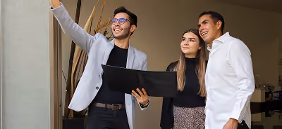 Un agent immobilier souriant tenant un dossier noir pointe vers le haut tout en faisant visiter une propriété à un jeune couple heureux.
