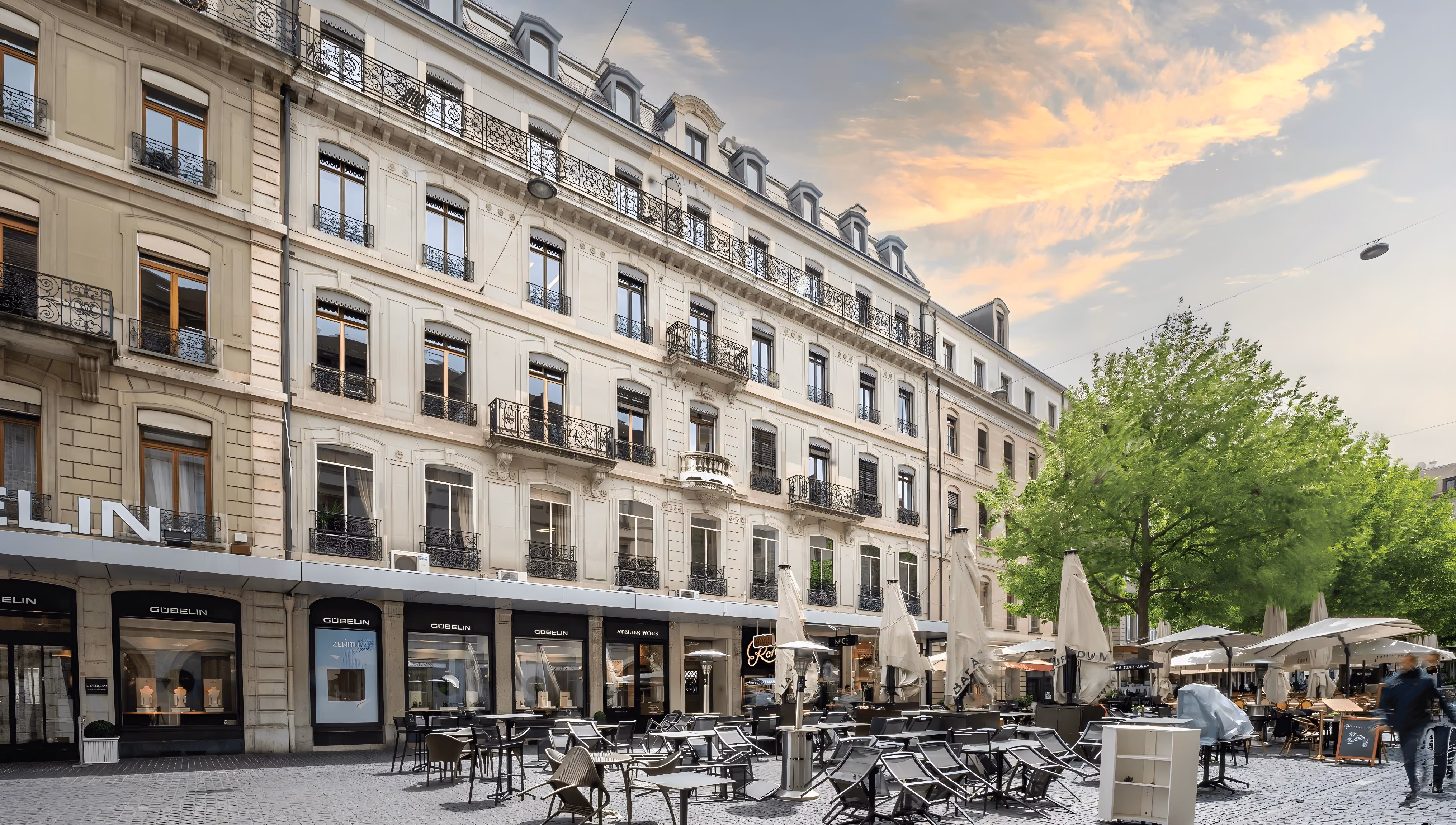 La place pavée du Molard à Genève, avec des boutiques de montres de luxe au rez-de-chaussée d'un bâtiment élégant et la terrasse d'un café avec des parasols.