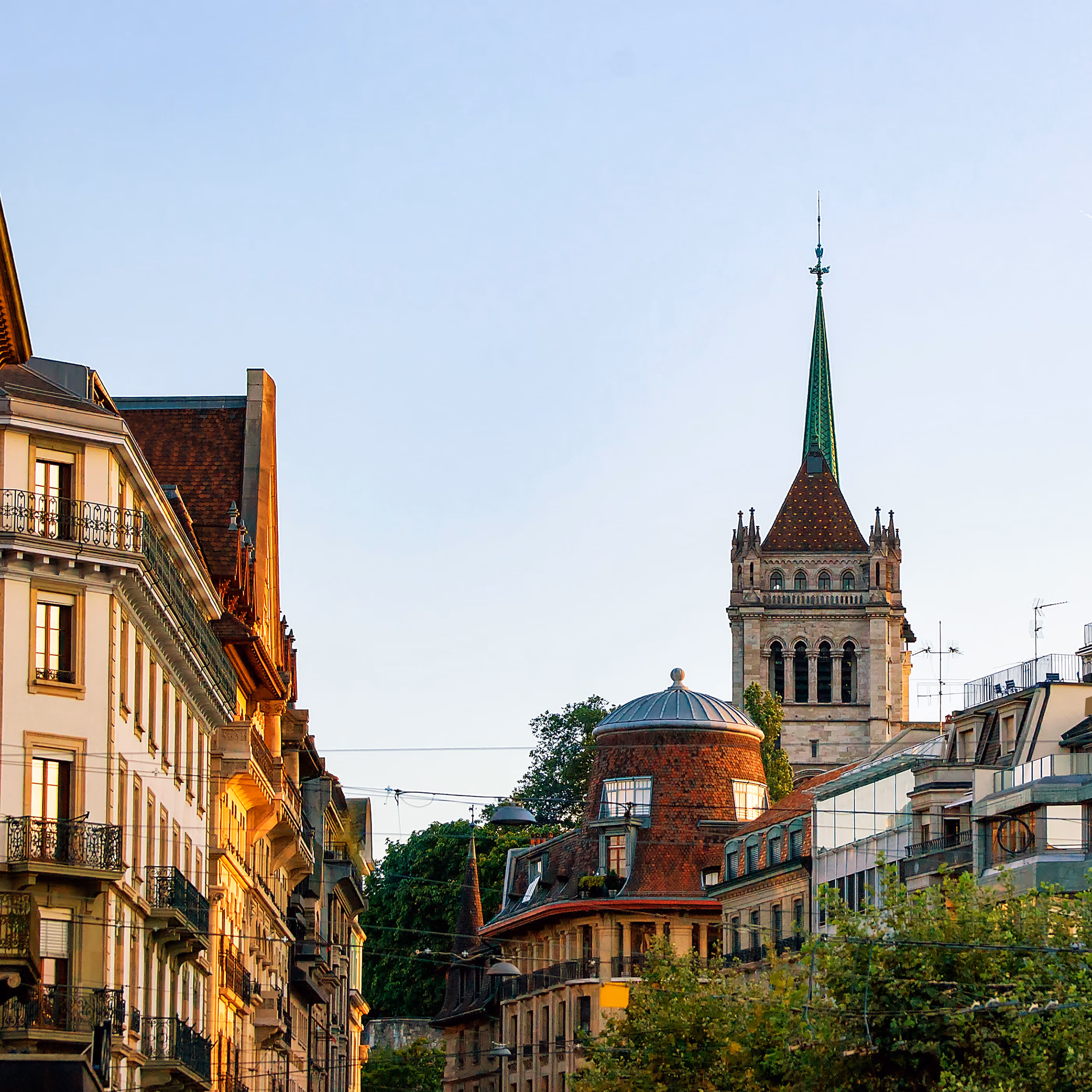 Une vue de rue de bâtiments classiques européens baignés par la lumière chaude du soleil, avec la flèche verte caractéristique et la tour en pierre de la cathédrale Saint-Pierre de Genève s'élevant en arrière-plan.