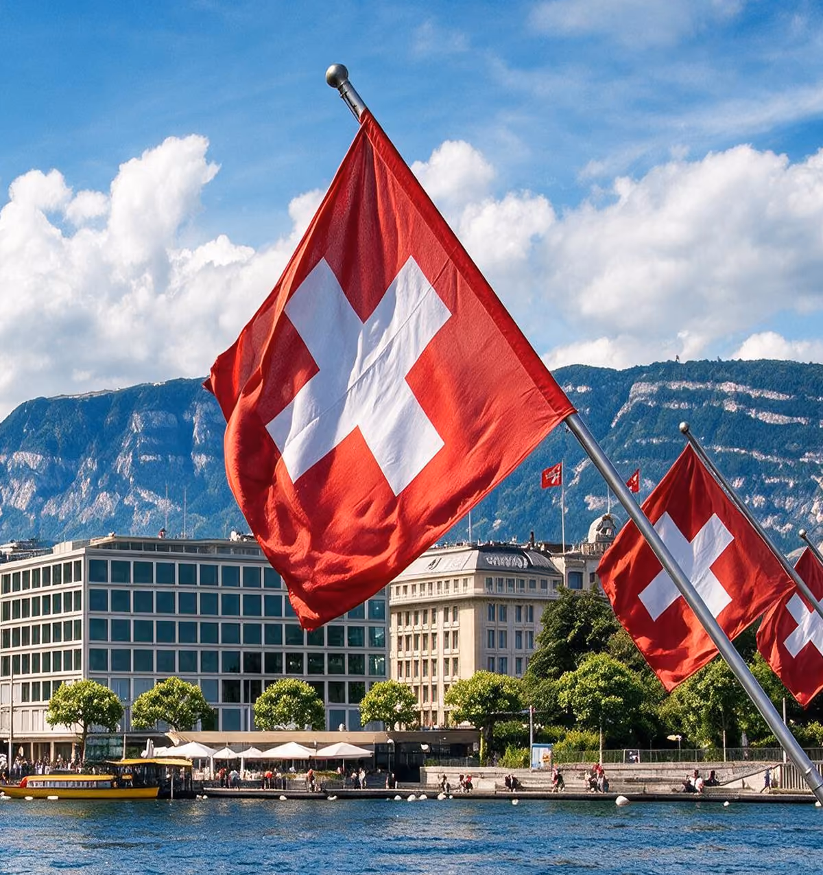 Des drapeaux suisses éclatants flottent bien en vue le long de la rive ensoleillée du lac Léman, avec des bâtiments au bord de l'eau, un bateau-taxi jaune et le mont Salève visible en arrière-plan.