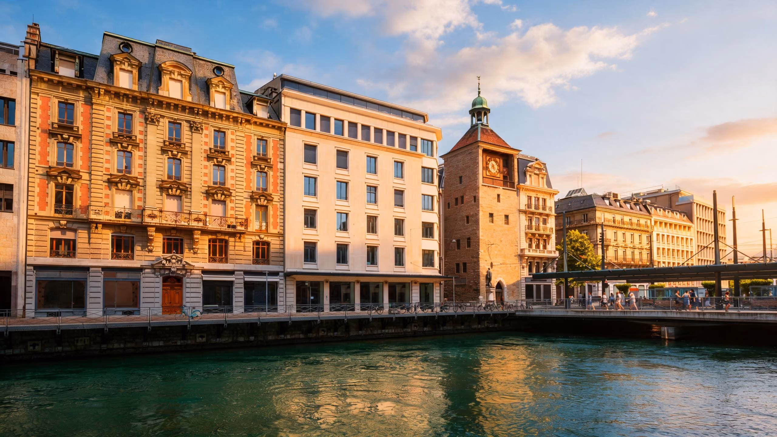 Des bâtiments historiques et une tour d'horloge en pierre baignés par la chaude lumière du coucher de soleil le long d'un quai à Genève.
