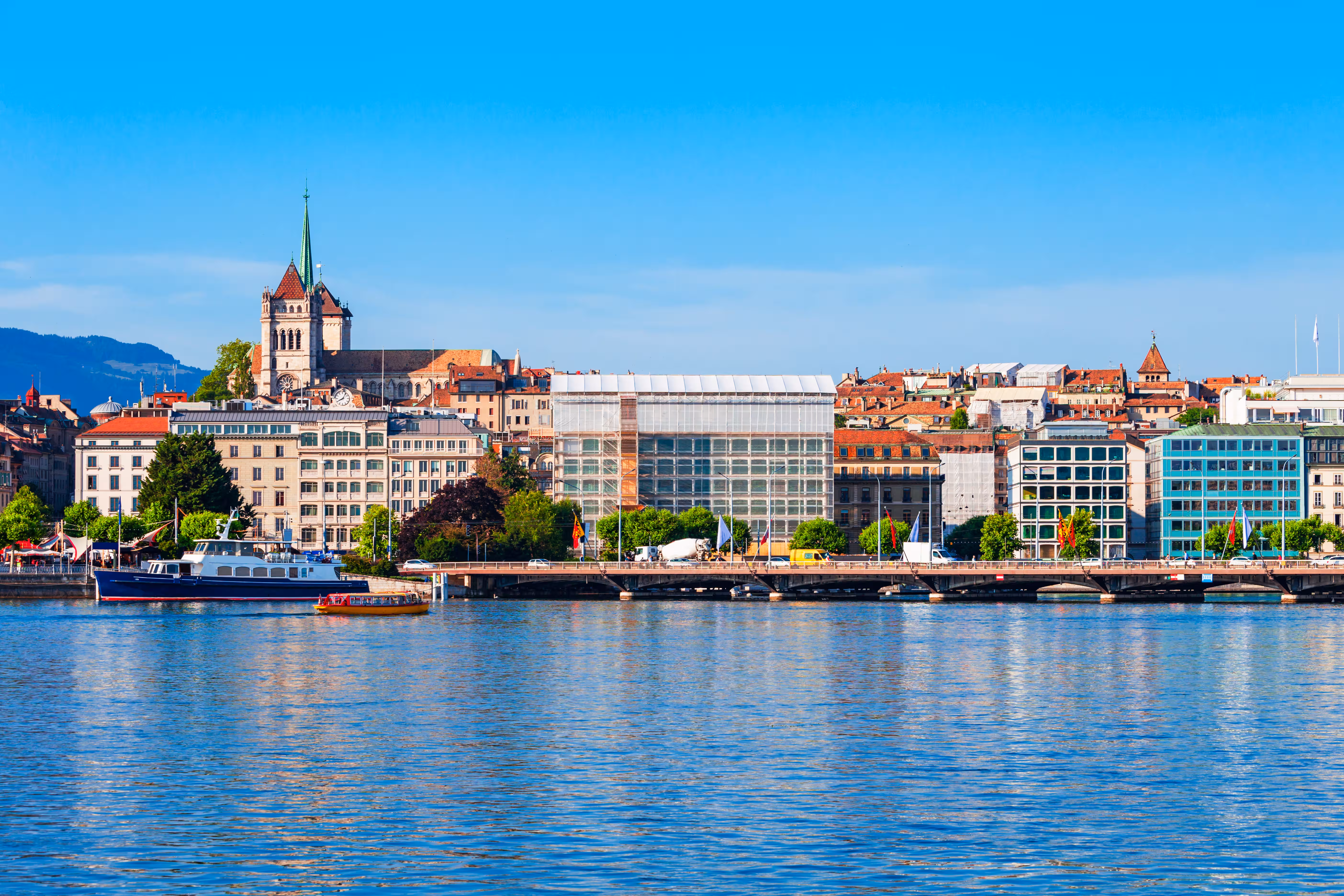 Une vue sur le lac Léman vers le quai et la vieille ville historique de Genève, avec la cathédrale Saint-Pierre s'élevant sur la colline, des bateaux naviguant sur l'eau bleue et un pont s'étendant le long de la rive.