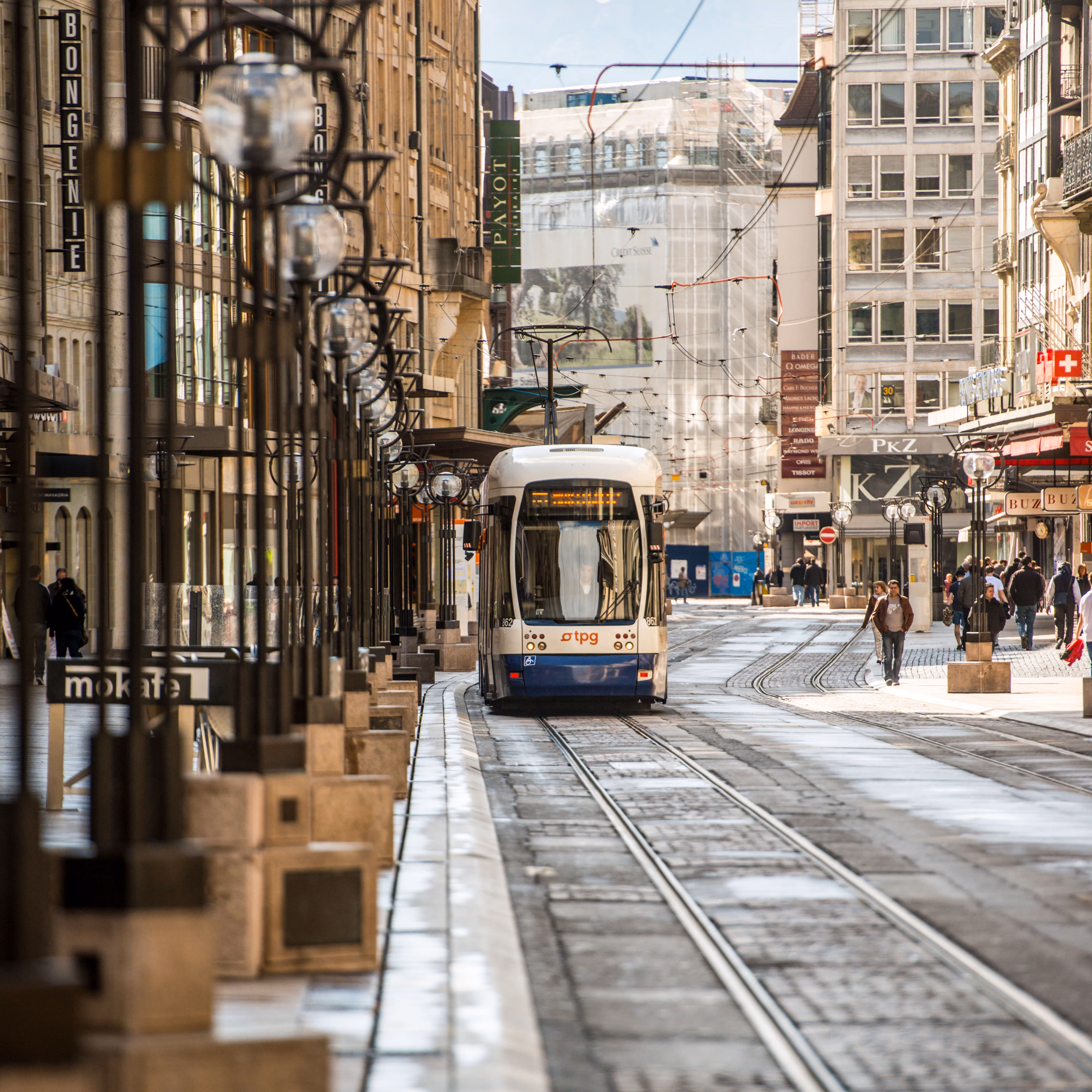 Un tramway TPG blanc et bleu circulant dans une rue commerçante animée de Genève, bordée de magasins, de piétons et de lampadaires modernes.