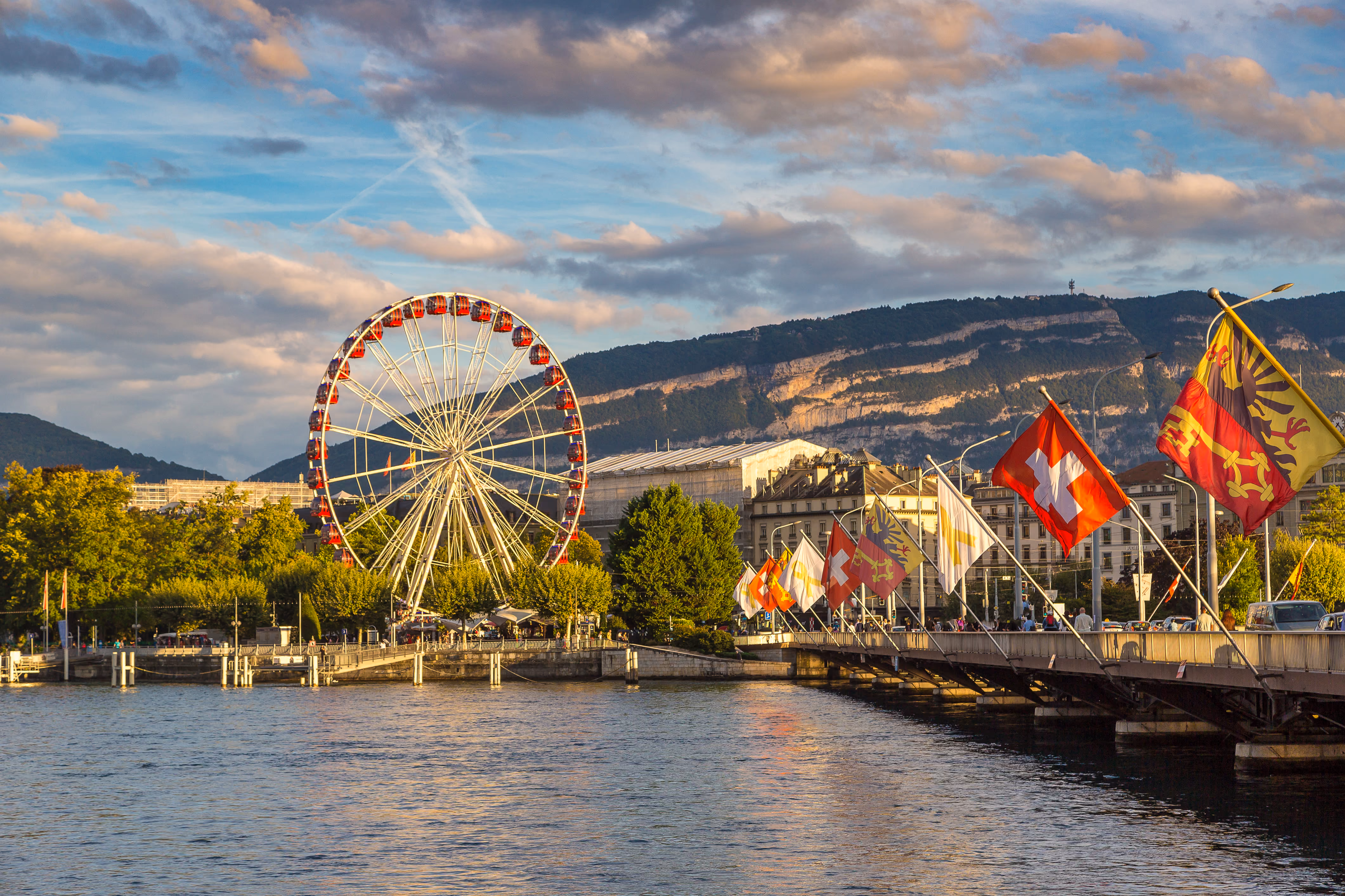 Une vue pittoresque de Genève au coucher du soleil, avec une grande roue sur les rives du lac Léman, le pont du Mont-Blanc bordé de drapeaux et le mont Salève illuminé en arrière-plan.
