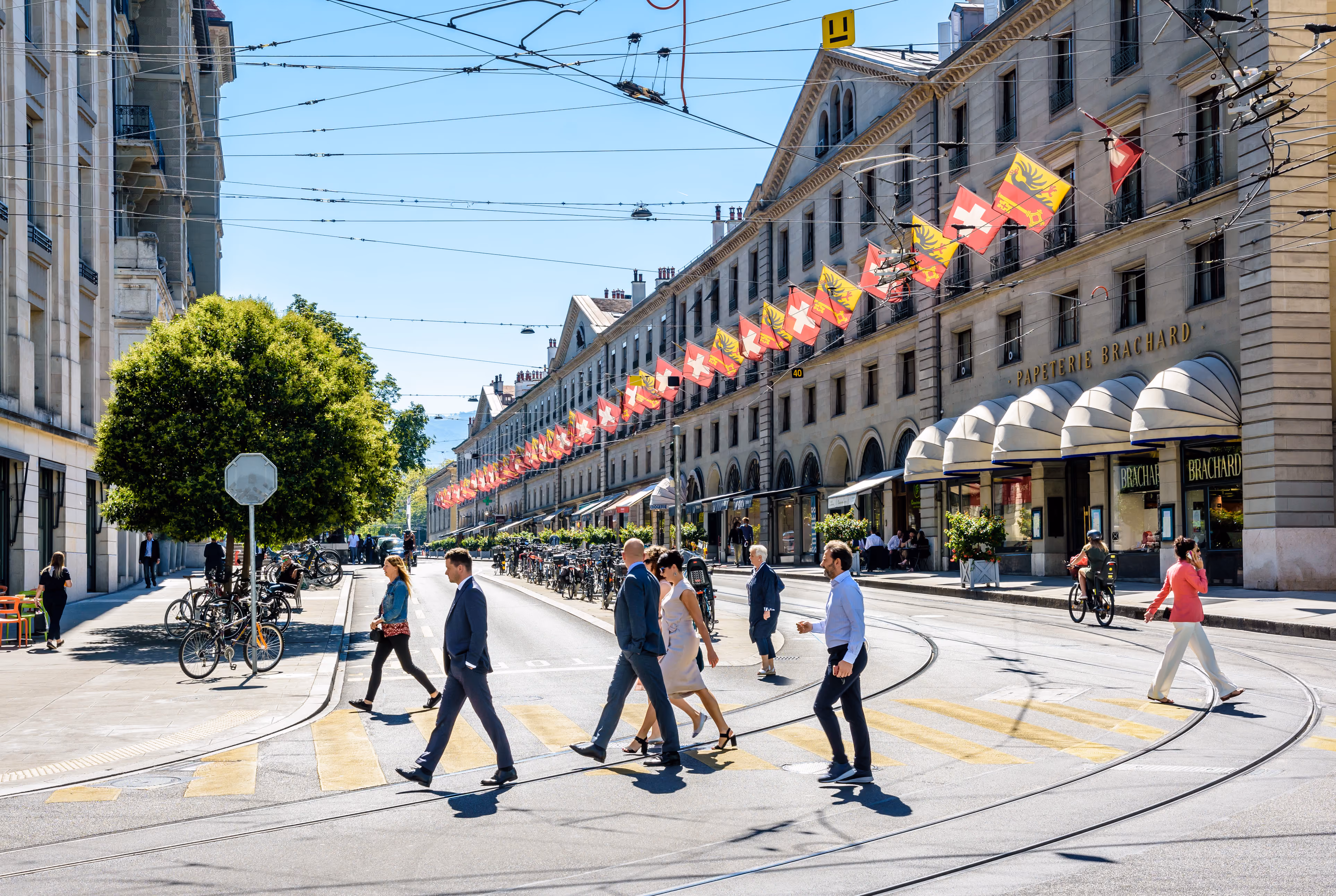 Des piétons traversant un passage piéton sur des voies de tramway à Genève, avec des drapeaux suisses et genevois suspendus entre les bâtiments historiques au-dessus de la rue.