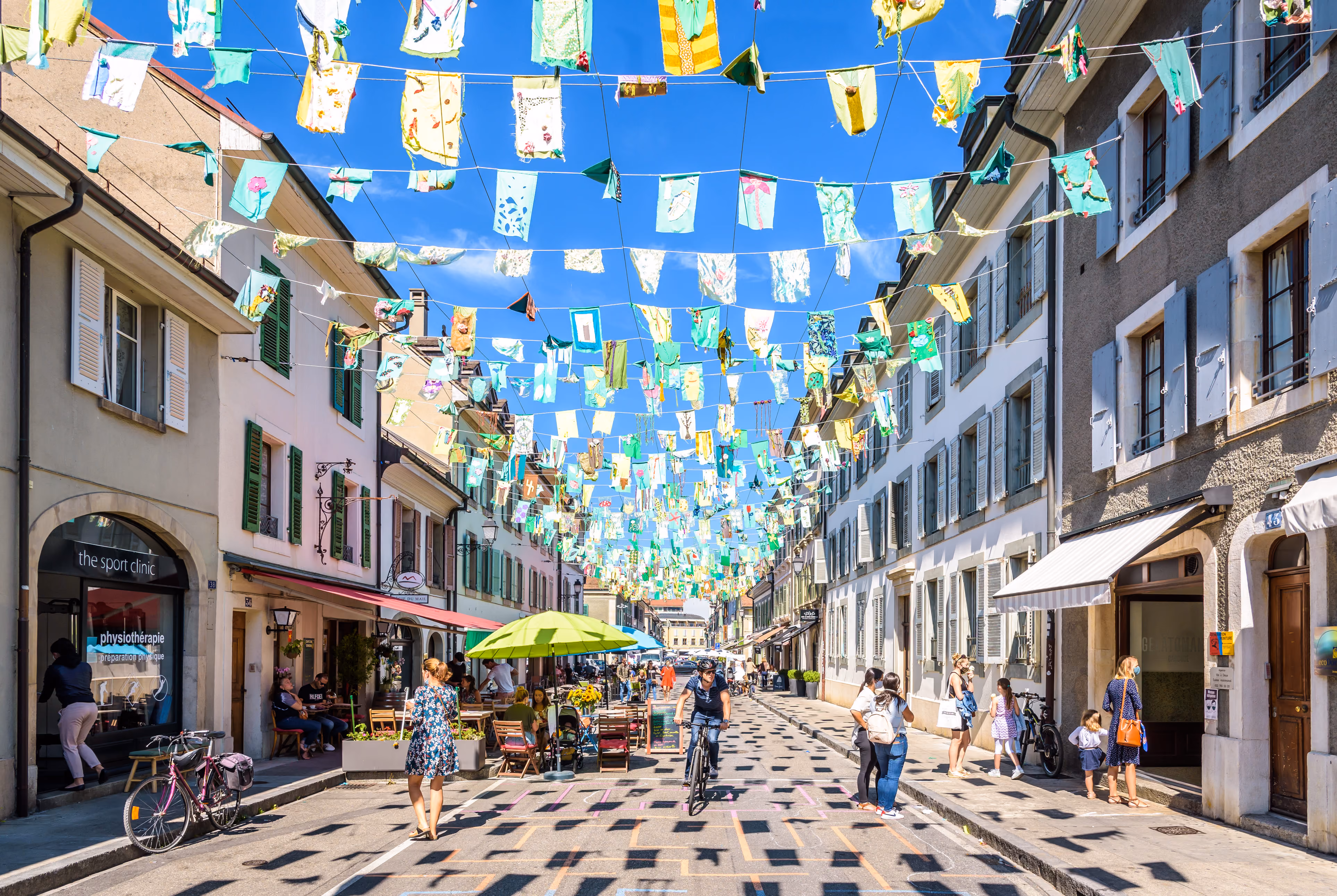 Une rue piétonne animée surplombée de guirlandes de fanions décoratifs colorés, avec des piétons, des cyclistes et des terrasses de café.