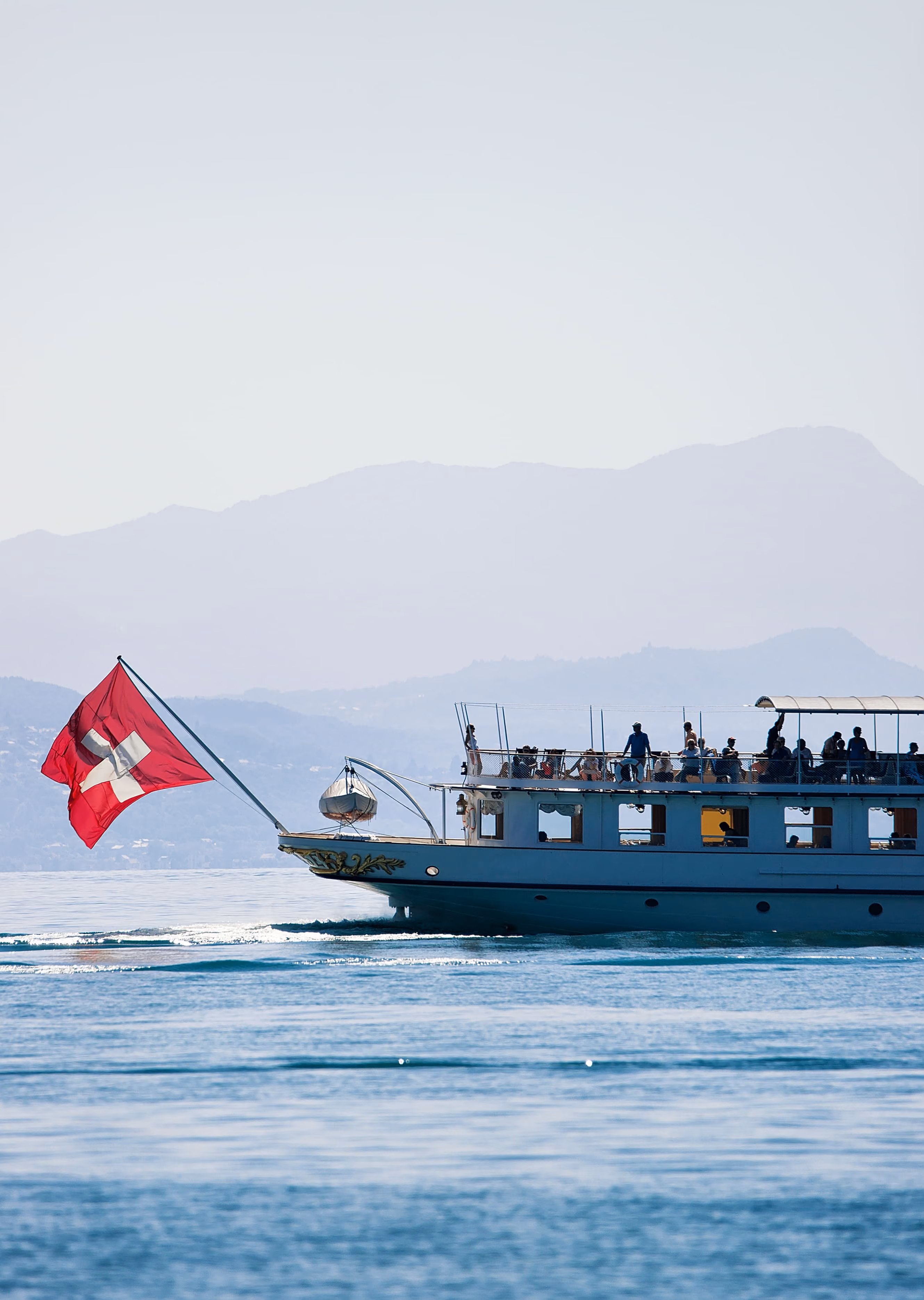 Un bateau de croisière arborant un grand drapeau suisse à la proue naviguant sur les eaux calmes d'un lac, avec la silhouette de montagnes au loin en arrière-plan.