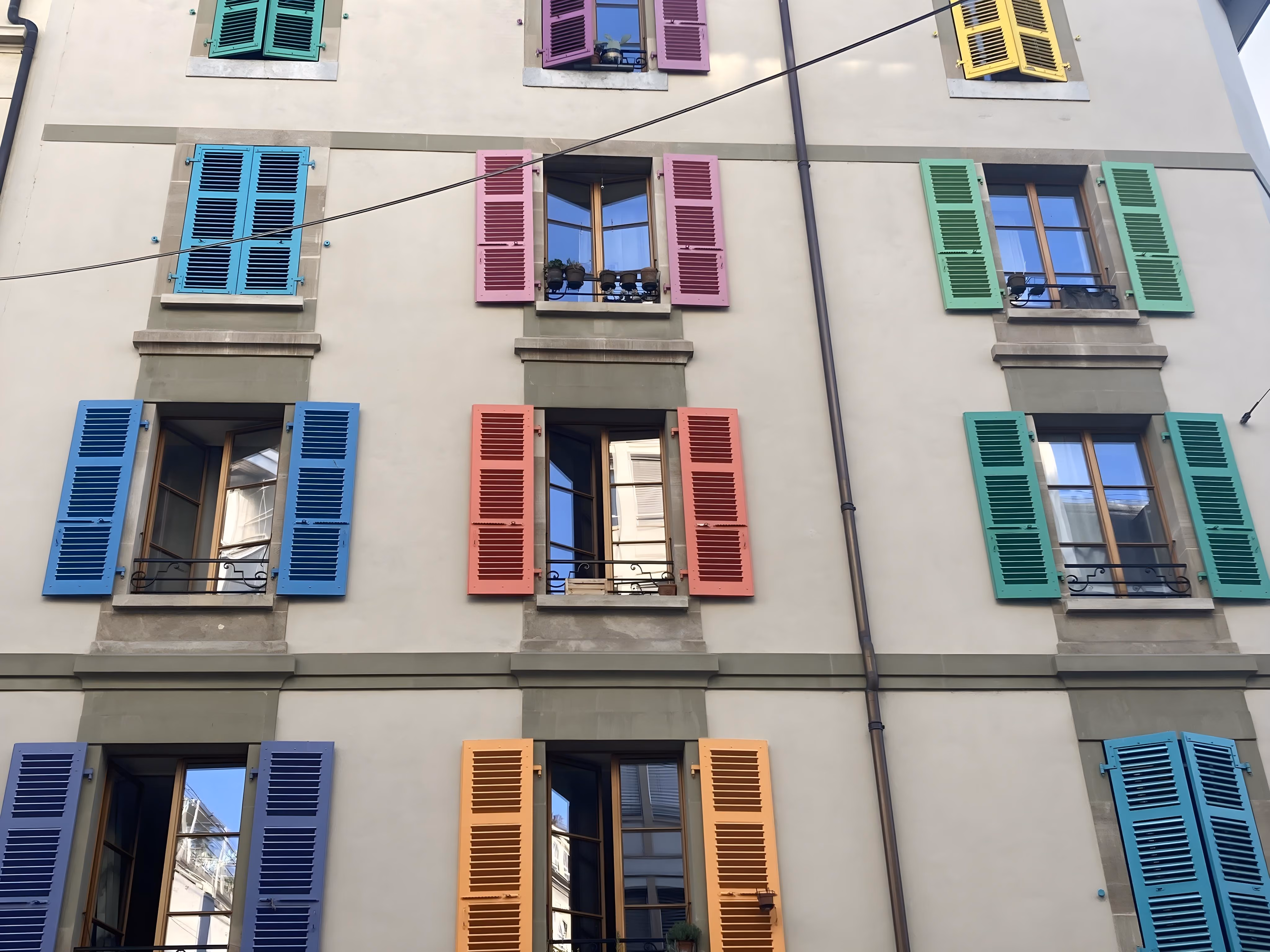 La façade d'un bâtiment présentant des rangées de fenêtres encadrées par des volets en bois multicolores éclatants aux nuances de vert, bleu, rose, orange et jaune.