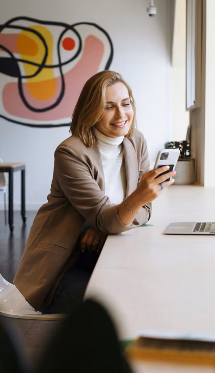 Smiling woman with blonde hair wearing a brown blazer and white turtleneck sits at a desk by a window, looking at her smartphone, with a laptop and abstract art on the wall behind her.