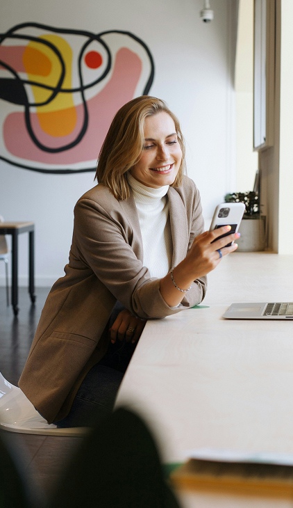 Smiling woman with blonde hair wearing a brown blazer and white turtleneck sits at a desk by a window, looking at her smartphone, with a laptop and abstract art on the wall behind her.