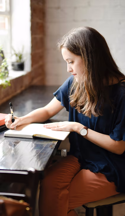 A woman with long brown hair wearing a navy blue shirt and orange pants is sitting at a table near a window, writing in an open notebook with a pen.