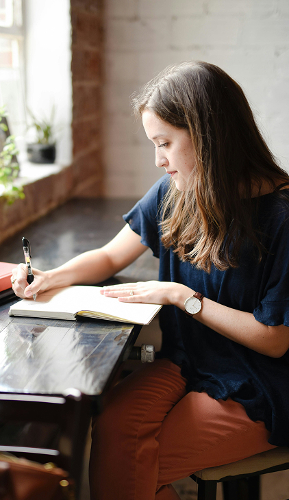 A woman with long brown hair wearing a navy blue shirt and orange pants is sitting at a table near a window, writing in an open notebook with a pen.