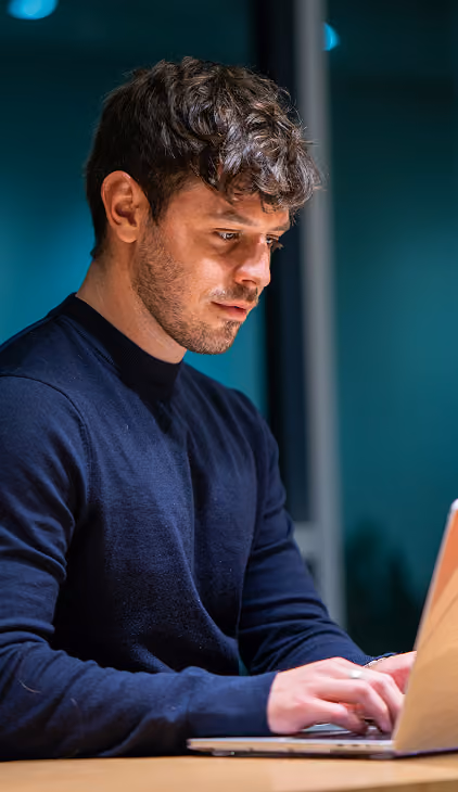 Young man with short curly hair and a beard, wearing a dark sweater, focused on typing on a laptop at a desk in a dimly lit room.