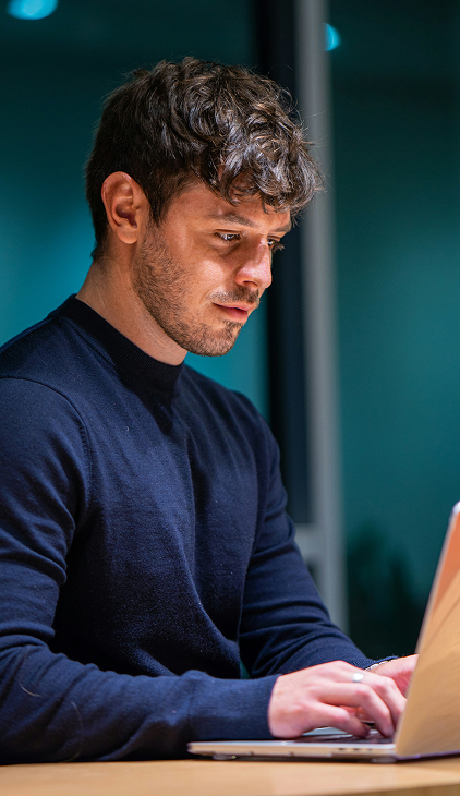 Young man with short curly hair and a beard, wearing a dark sweater, focused on typing on a laptop at a desk in a dimly lit room.