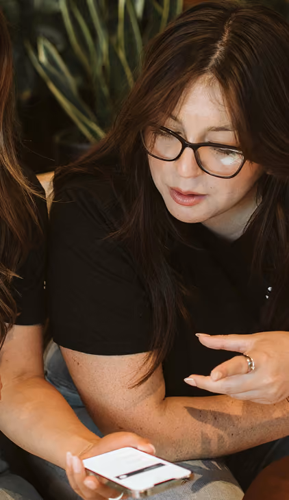 Woman with long dark hair and glasses wearing a black shirt looking down at a smartphone held by another person.