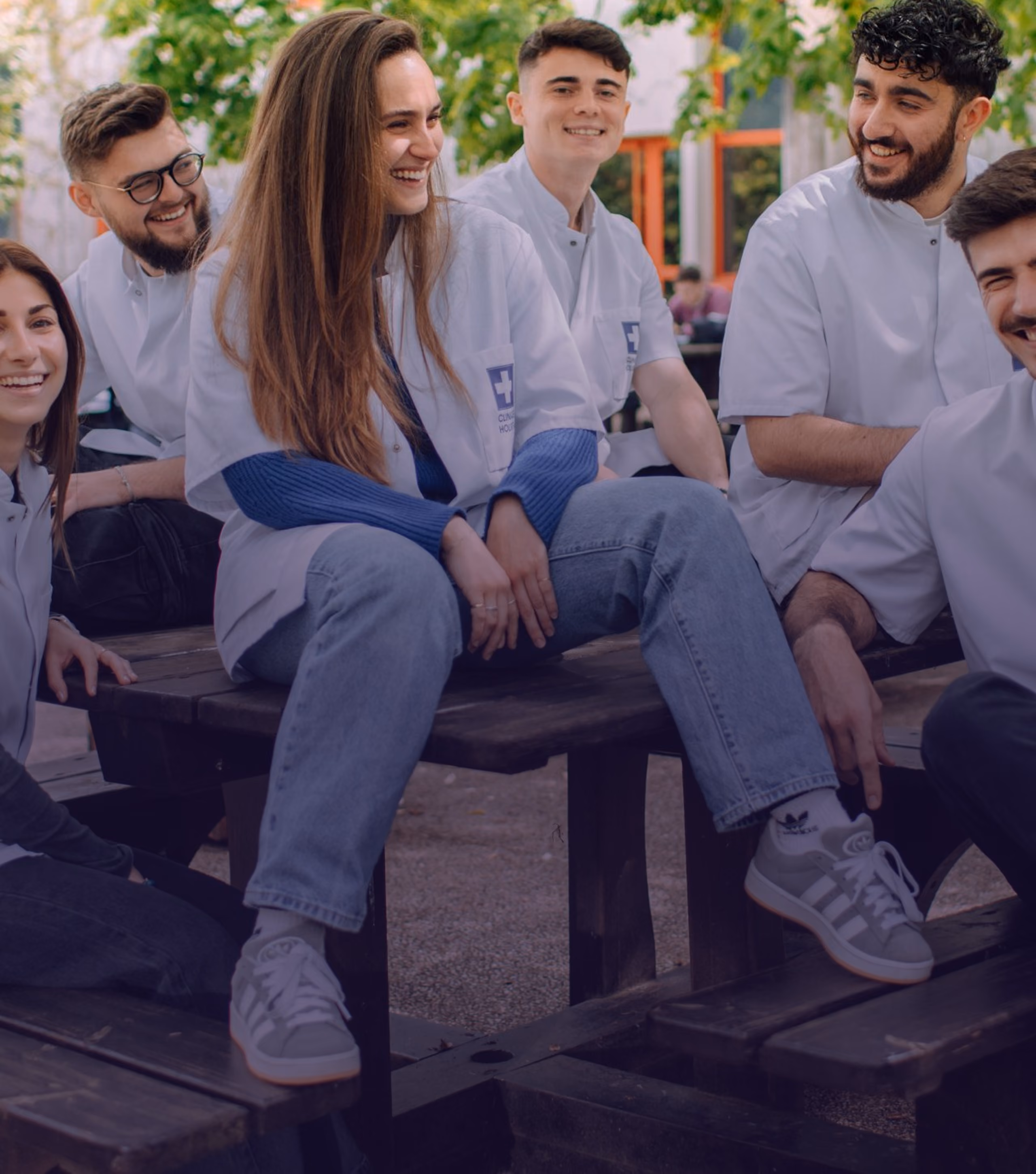 Group of six young adults wearing white lab coats sitting and smiling on wooden benches outdoors, with greenery and a building in the background.