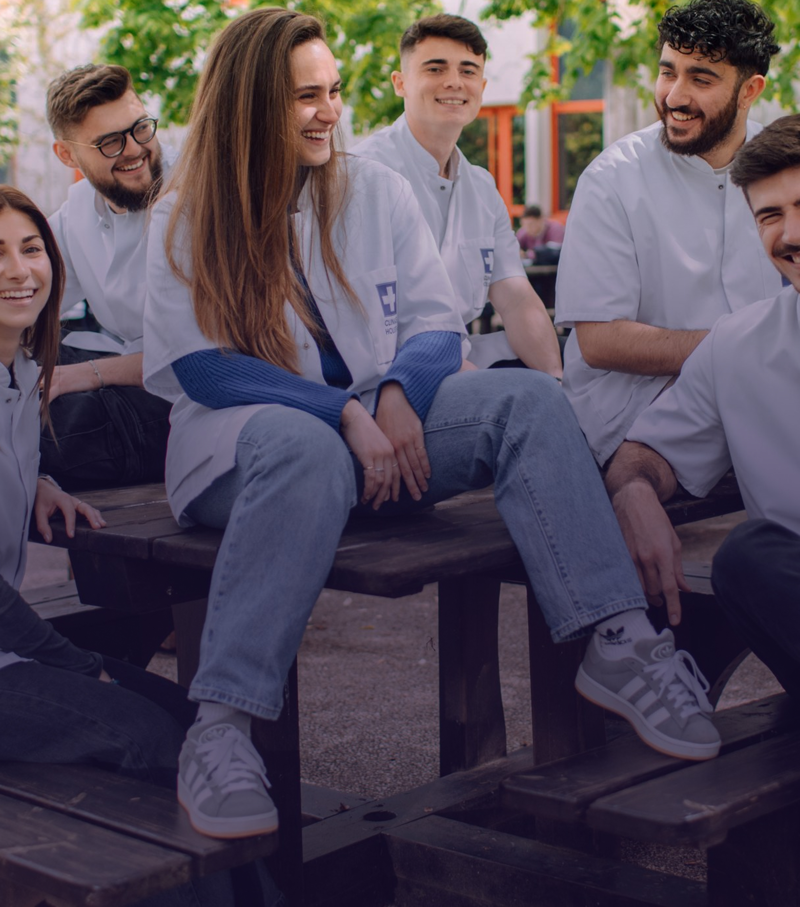 Group of six young adults wearing white lab coats sitting and smiling on wooden benches outdoors, with greenery and a building in the background.