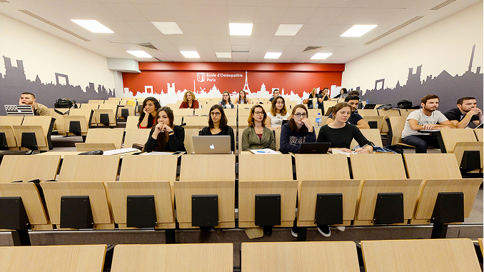 Students sitting in a lecture hall with tiered wooden seats, some using laptops, facing forward; the back wall is red with white silhouettes of Paris landmarks and the words Ecole d'Osteopathie Paris.