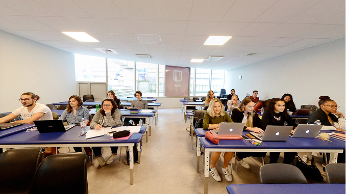Students seated in a bright classroom with multiple rows of blue tables, some using laptops and others taking notes, facing forward under fluorescent ceiling lights.