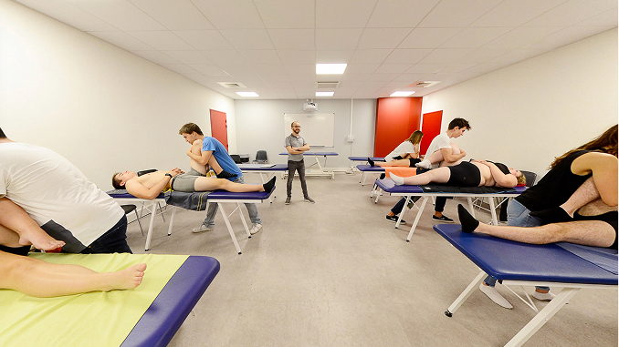 A group of people receiving physical therapy in a bright room with white walls and a gray floor; six therapy tables are arranged with pairs of therapists and patients, and a man stands in the center observing.