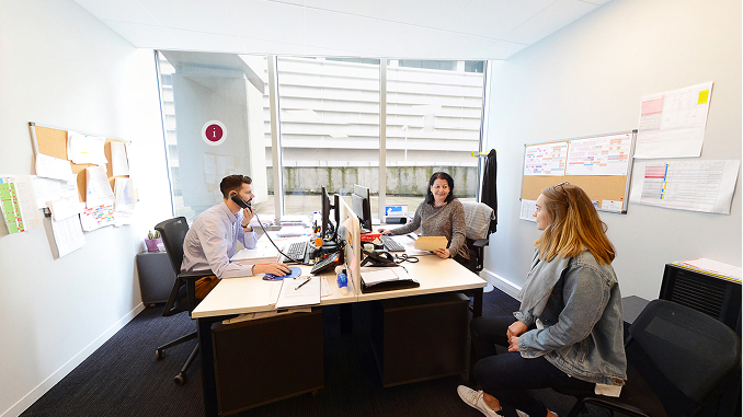 Three people in an office: a man wearing a face mask talks on the phone at a desk with two computer monitors, a woman with dark hair holds papers and smiles at him, and a blonde woman in a denim jacket sits nearby on a chair.