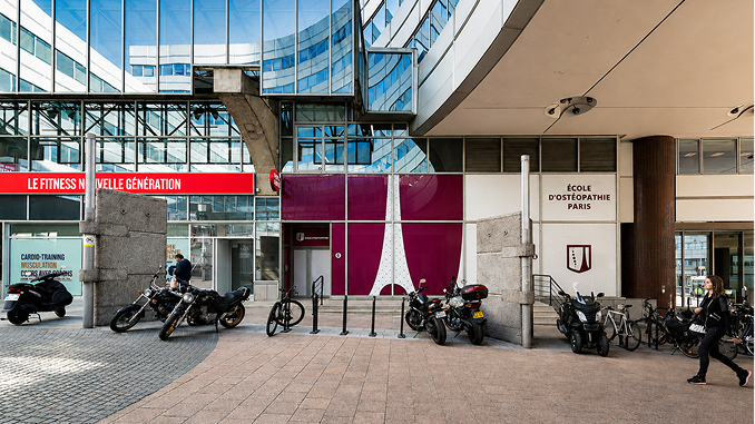 Entrance of École d'Ostéopathie Paris with motorcycles and bicycles parked outside under a modern glass and concrete building facade, and a person walking on the right side.