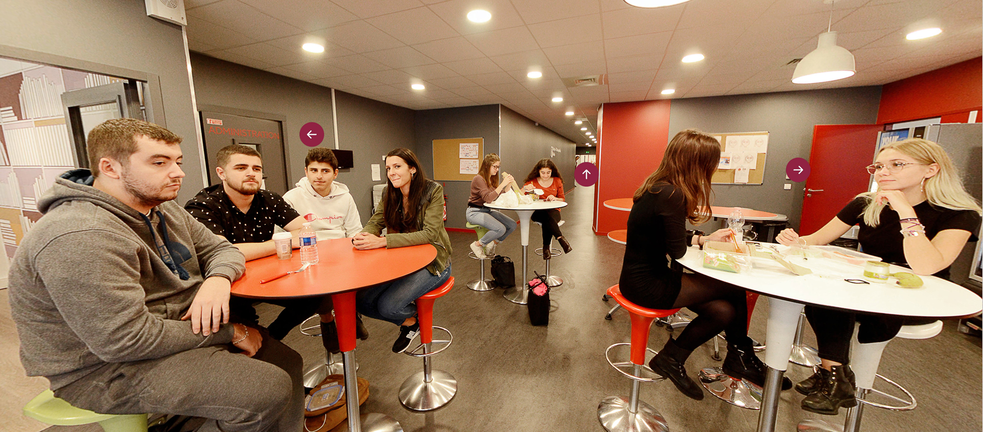 Seven young adults sitting at three tall tables in a modern indoor lounge area with gray walls and ceiling lights, some of them eating and chatting.