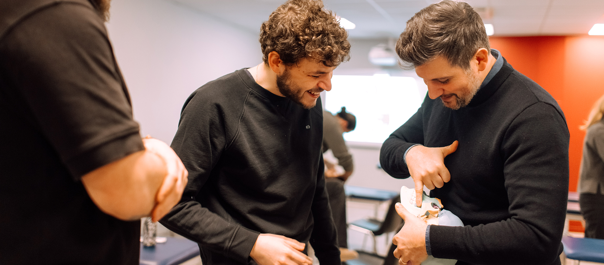 Two men smiling and examining a human skull model indoors, with one man pointing at the model's teeth.