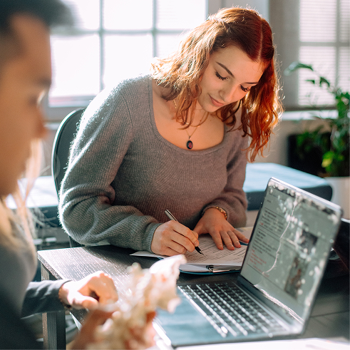 Young woman with red hair wearing a gray sweater writing on a document at a table with an open laptop while another person is nearby holding a seashell, in a bright room with large windows.