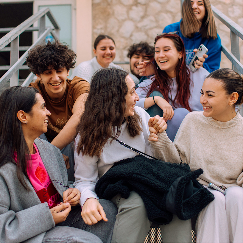 A group of seven young people sitting on stairs outdoors, smiling and interacting happily, with one pair holding hands in the front.