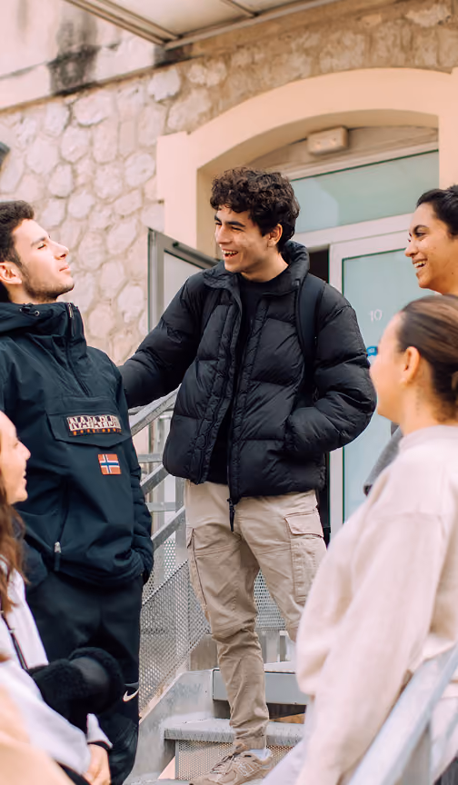 A group of five young adults standing outdoors on steps next to a building, smiling and engaging in conversation, dressed in winter jackets and casual clothes.