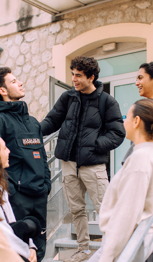 A group of five young adults standing outdoors on steps next to a building, smiling and engaging in conversation, dressed in winter jackets and casual clothes.