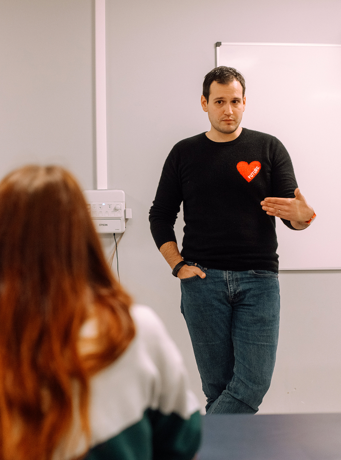 A man in a black sweater with a red heart and the word 'FUTURE' on it is standing and gesturing during a conversation or presentation, facing a seated person with long reddish hair in a white and green sweater, in a room with a whiteboard on the wall.
