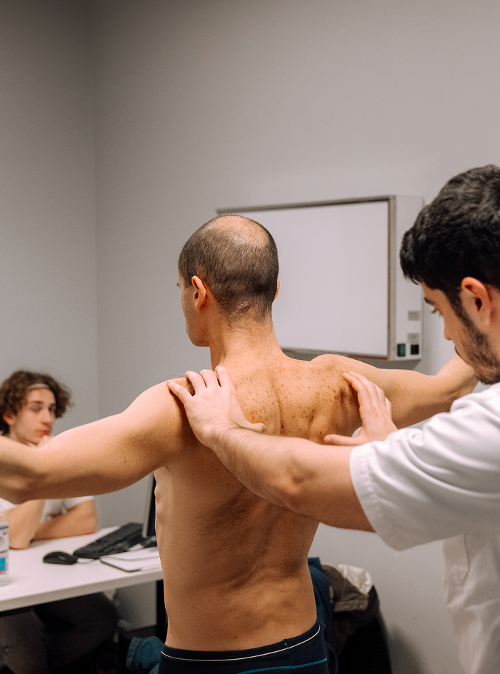 A healthcare professional examining the back and shoulder movement of a shirtless man standing with his arms extended in a clinical setting, while another person watches from a desk in the background.