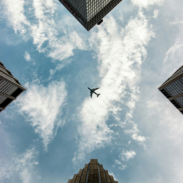 View looking up at a blue sky with scattered clouds framed by four tall city buildings with a plane flying in the center.