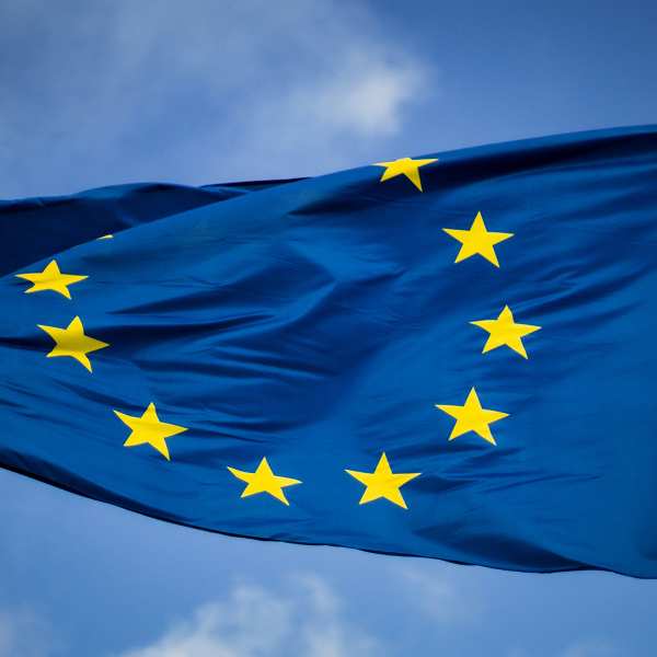 European Union flag with twelve yellow stars in a circle on a blue background waving against a partly cloudy sky.